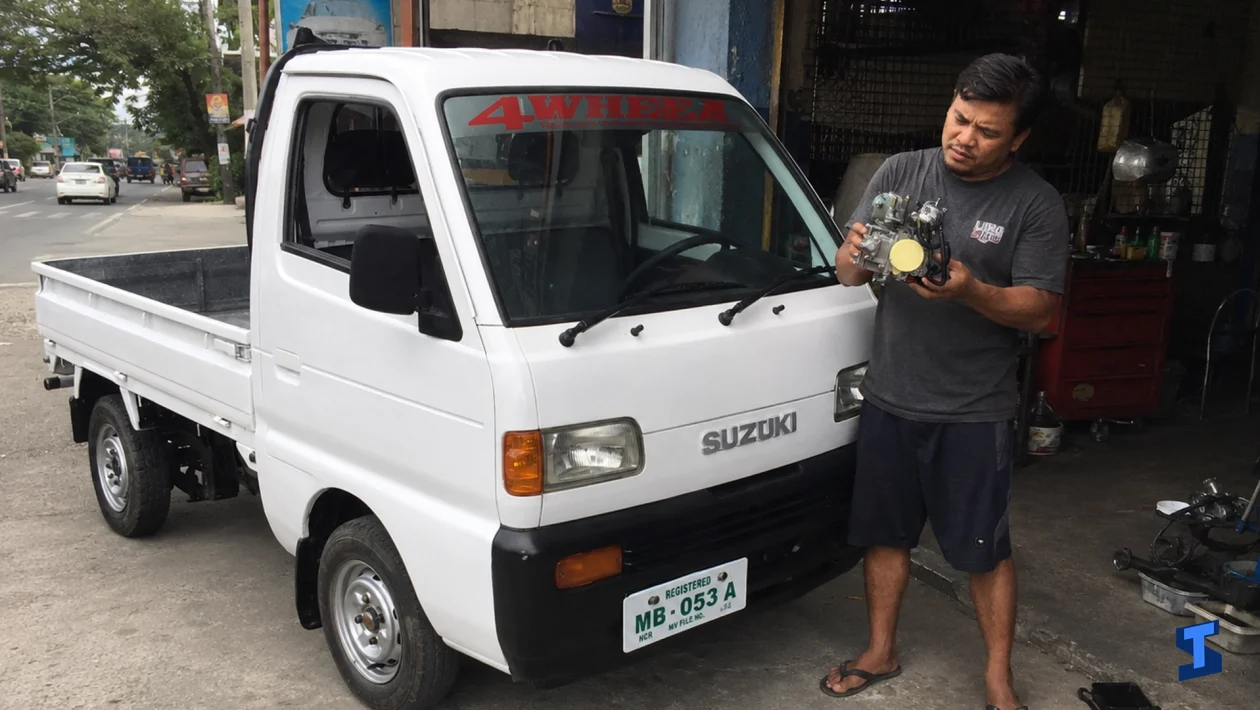 A man stands beside a white Suzuki multicab outside a roadside car shop while holding a carburetor and inspecting it near the front of the truck.