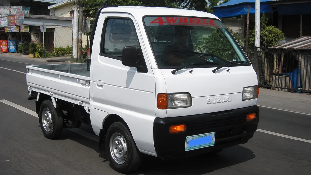 A white Suzuki kei truck driving on a paved Philippine road in daylight, with roadside shops, utility poles, overhead wires, trees, and local buildings in the background.