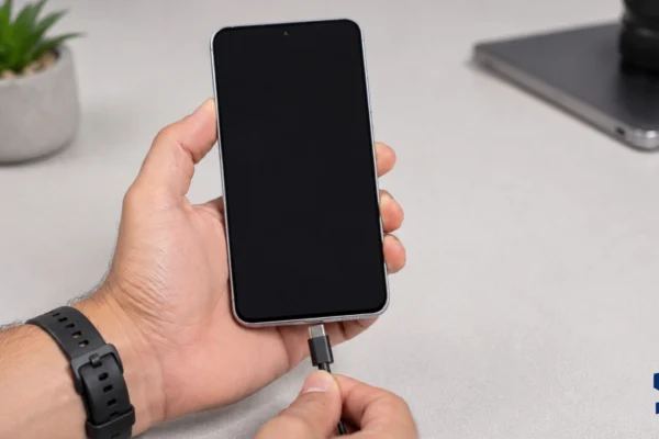 Samsung Galaxy A57 held in one hand while a user connects a USB-C charging cable to the phone, showing a black screen on a clean table with a blurred plant in the background.